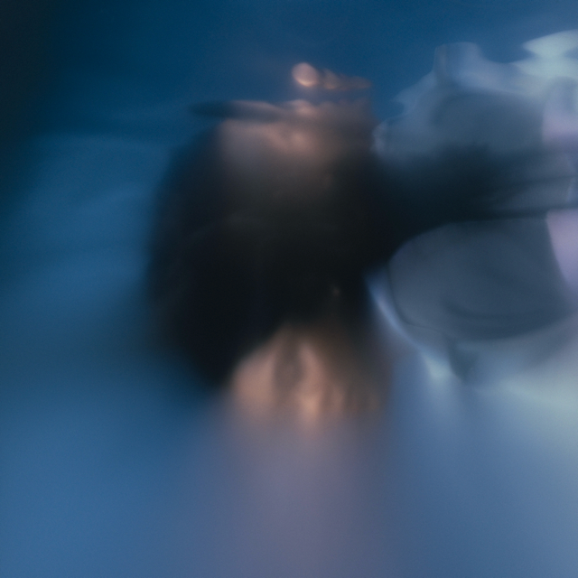 An abstract, heavily blurred photograph with indistinct human-like forms and soft blue, white, and dark tones, resembling a dreamlike or motion-blurred artistic scene.
